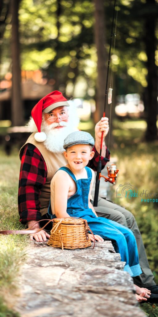 Santa Claus wearing a vest, a red and black plaid shirt and holding a fishing pole. A young boy sits in front of him wearing blue overalls.