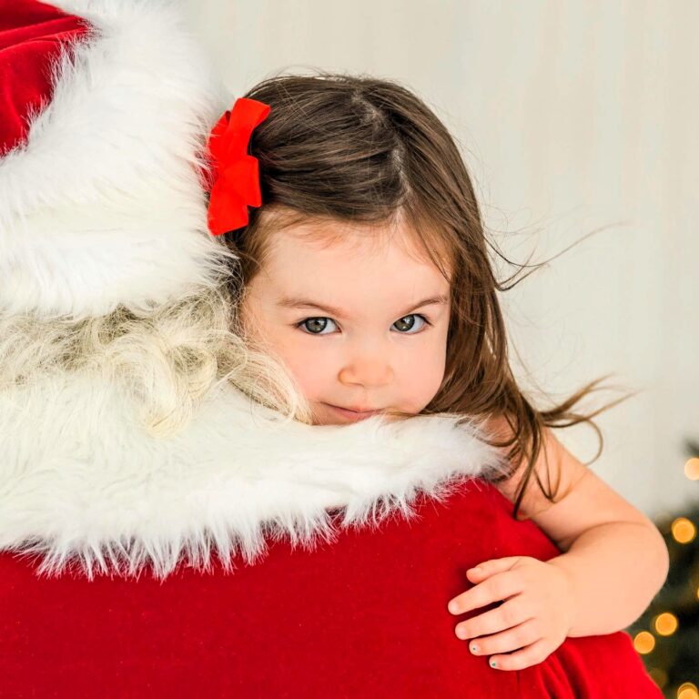 A small girl with brown hair is hugging Santa Claus and looking at the camera.