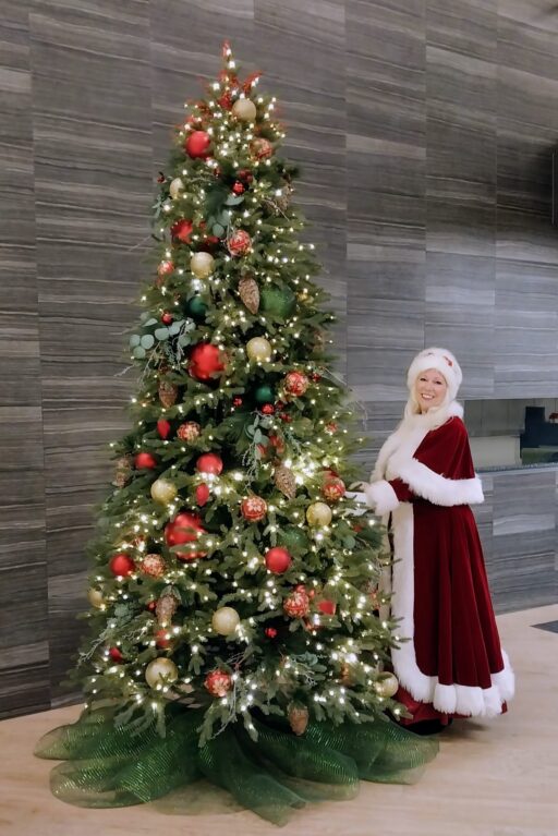 Mrs. Claus wearing a long red robe and white fur hat, standing next to a tall Christmas tree.