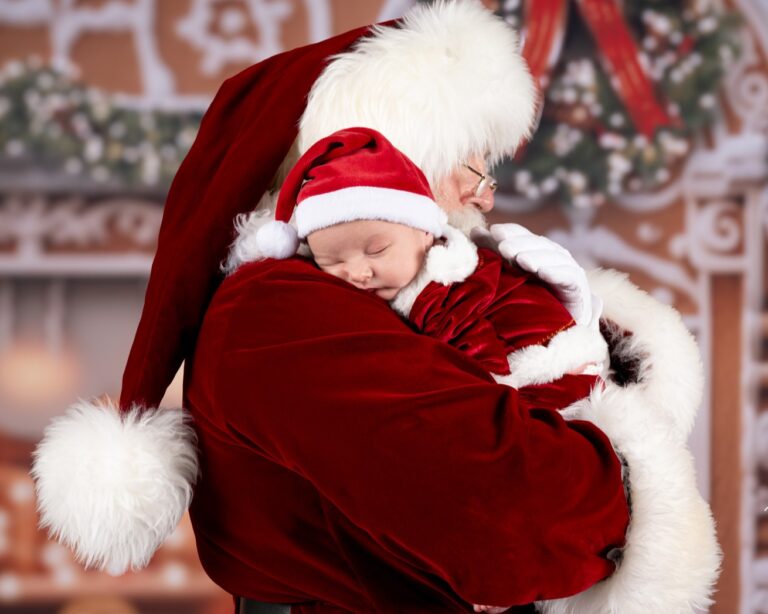 Santa Claus is holding a two week old baby who is also wearing a Santa suit. The baby is sleeping on Santa's shoulder.