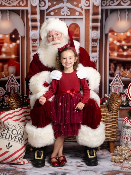Santa Claus sitting in a chair with a little girl standing in front of him. Both are looking at the camera.