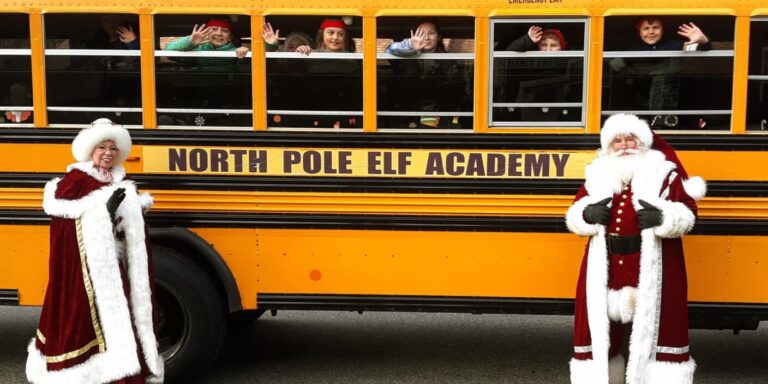 Santa and Mrs. Claus standing beside an orange school bus bearing a sign, "NORTH POLE ELF ACADEMY". Children are waving from the windows.