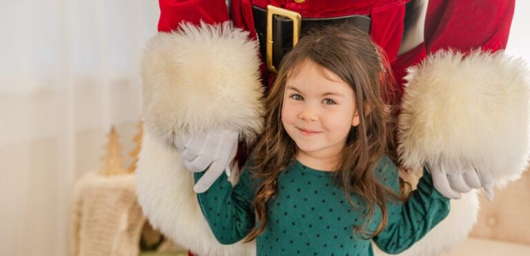 A young girl with brown hair is holding Santa Claus's hands and smiling at the camera.