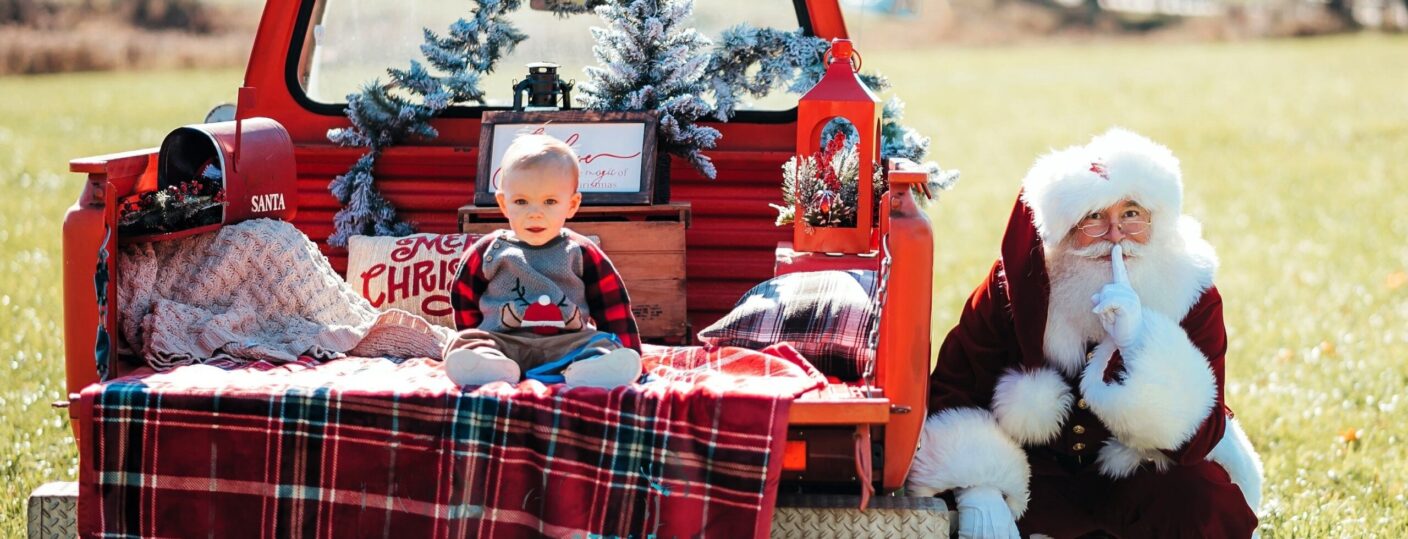 Santa looks at the camera with his finger in front of his mouth motioning for quiet as a 10 month old boy sits on the tailgate of a red pickup truck.