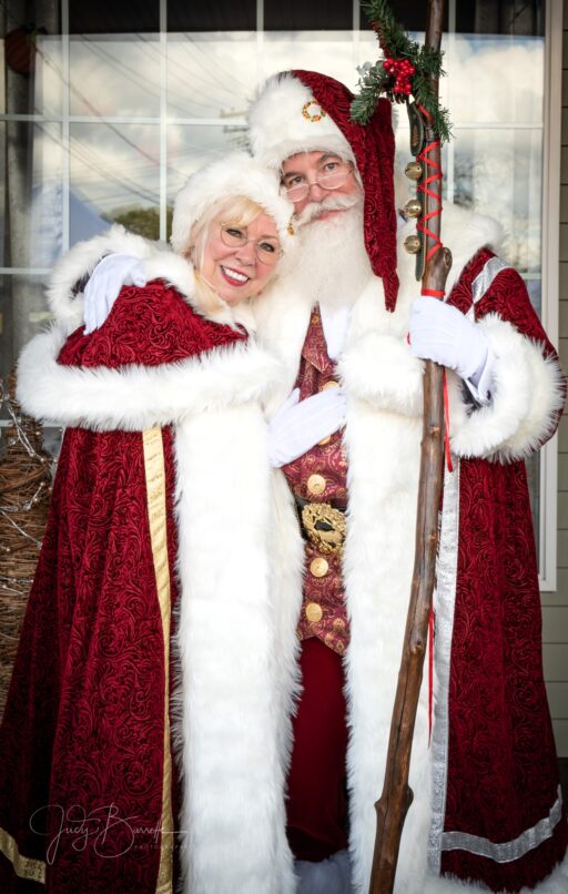 Santa and Mrs. Claus wearing long robes and standing in front of a shop window.