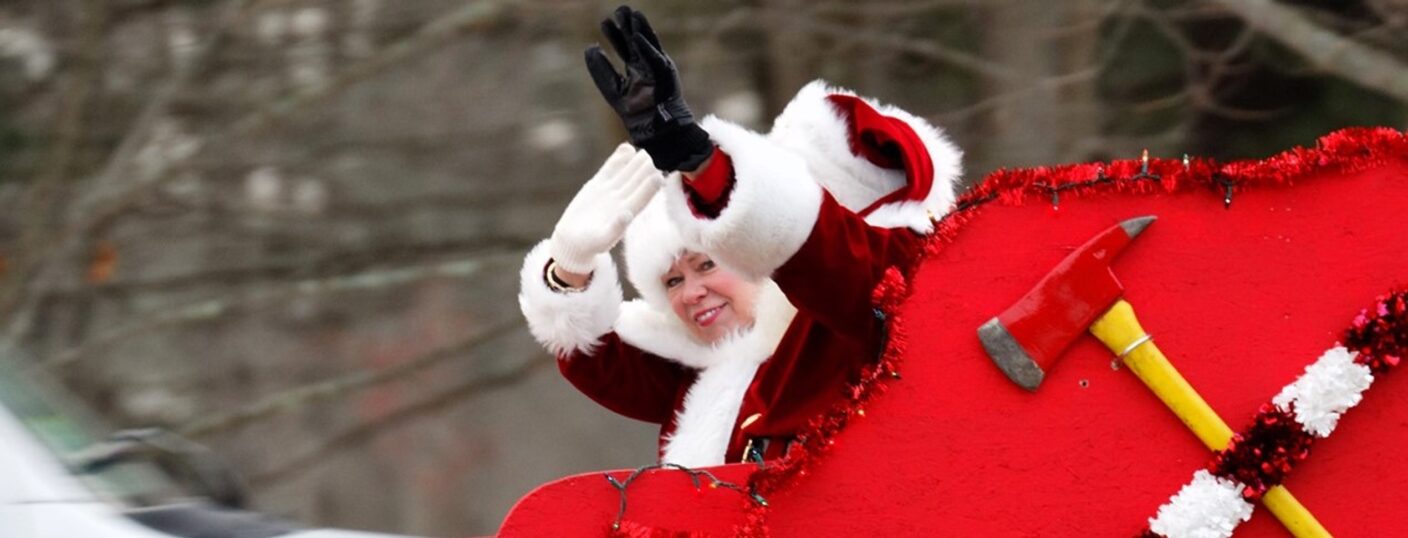 Mrs. Claus waiving at the camera while riding in a fire department Christmas sleigh.