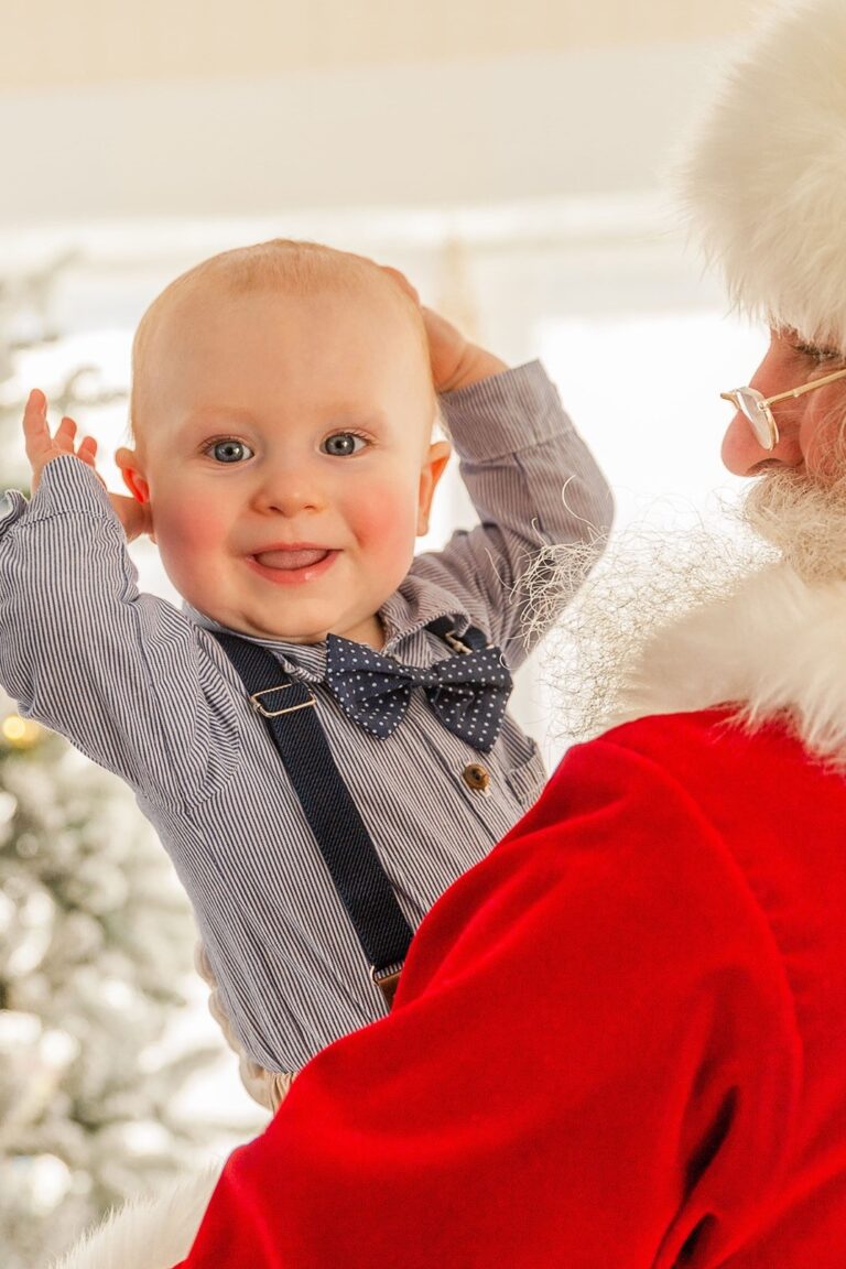 Santa Claus is holding a six-month old baby boy who is wearing suspenders and a bow tie, smiling at the camera with his hands raised.