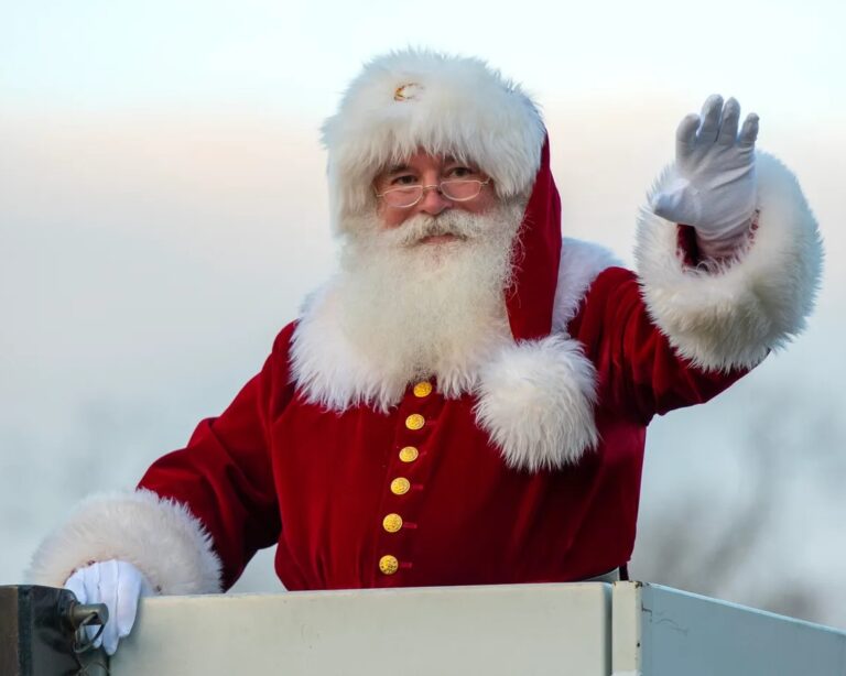 Santa Claus is standing in the bucket of a fire truck and waving at an unseen crowd.