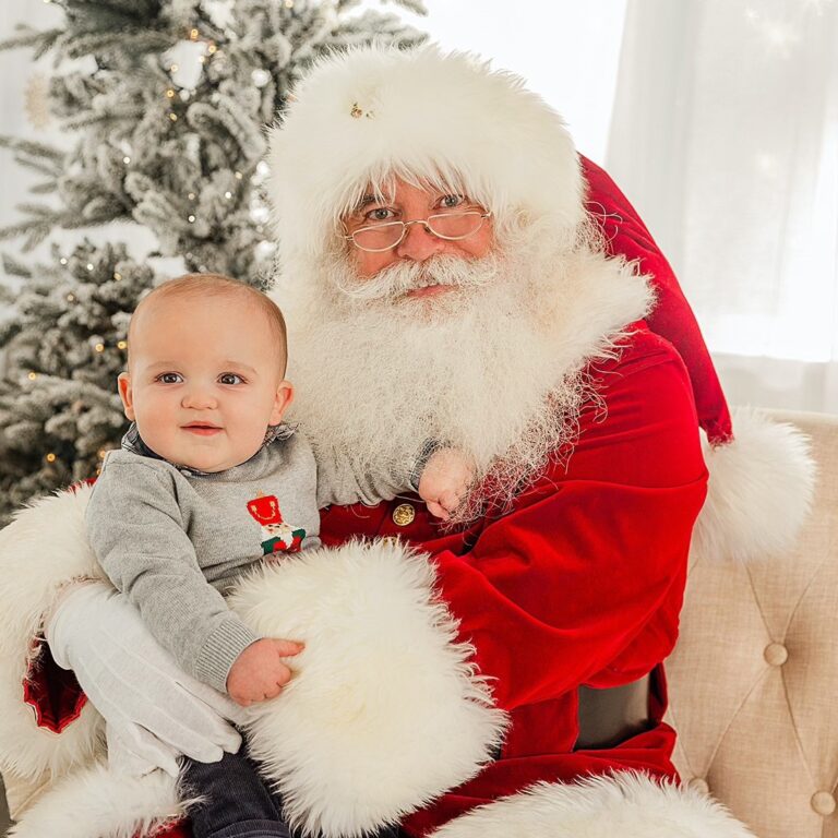 Santa Claus is sitting with a six-month old baby and smiling at the camera.
