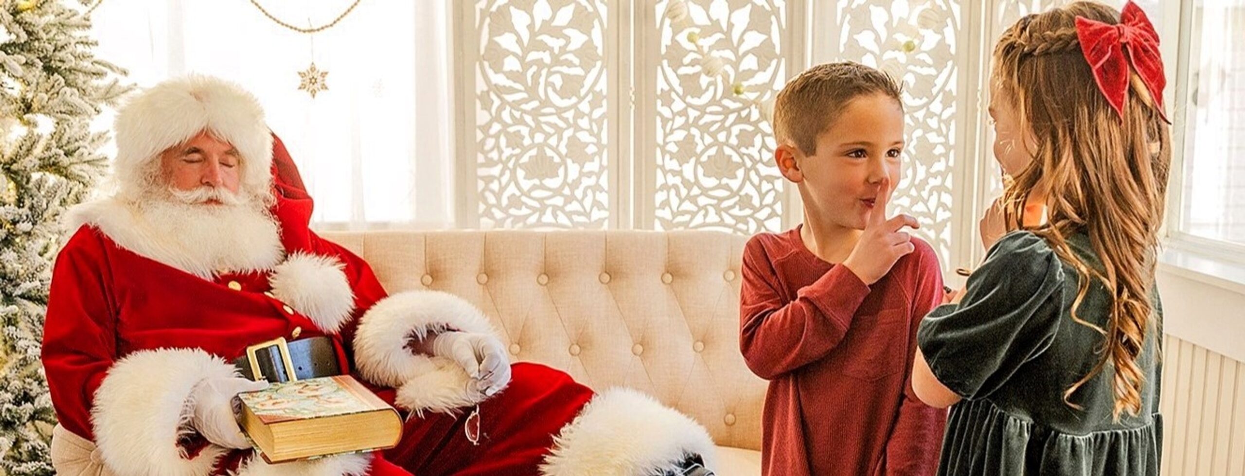 Santa Claus is resting with his eyes closes as two children look at him. The boy is motioning for his sister to be quiet.